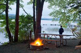 Ein Mann blickt auf den See, im Vordergrund lodert ein Feuer an einem Campingplatz