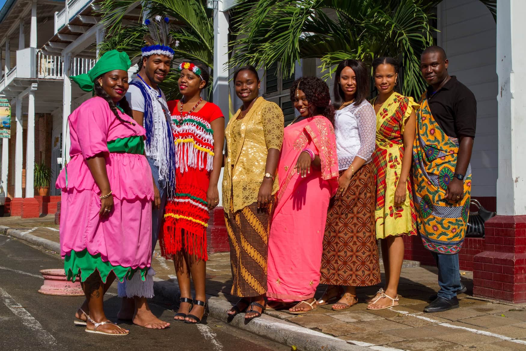 Junge Menschen in traditioneller Kleidung verschiedener Bevölkerungsgruppen Surinames vor Holzhäusern in Paramaribo.