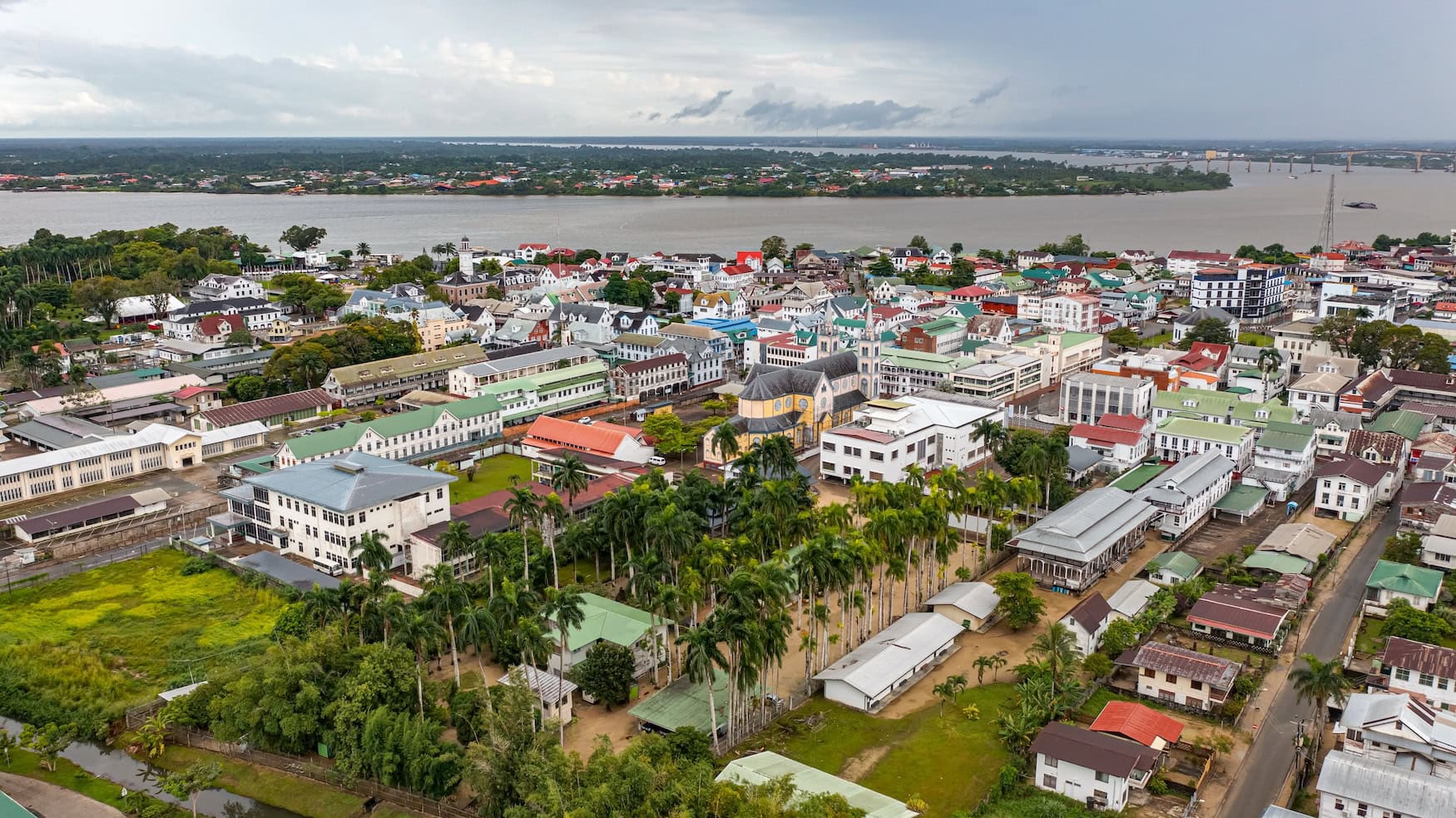Drohnenaufnahme der historischen Innenstadt von Paramaribo, mit Holzhäusern aus der niederländischen Kolonialzeit entlang der Uferpromenade des Suriname-Flusses unter einem wolkigen Himmel.