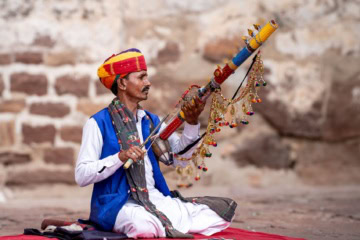 Rajasthanischer Musiker, der im farbenfrohen Turban und weißen Kurta-Pyjama-Lokalkleidung vor einer Ziegelmauer sitzt