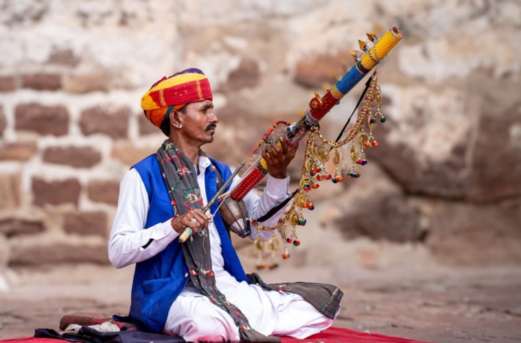 Rajasthanischer Musiker, der im farbenfrohen Turban und weißen Kurta-Pyjama-Lokalkleidung vor einer Ziegelmauer sitzt
