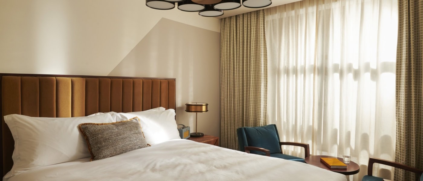 Ein Hotelzimmer im Mid-Century-Stil, mit breitem Bett, mit Samt gepolstertem Kopfteil und einer schwarzen Kunststoff-Designlampe an der Decke.