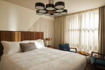 Ein Hotelzimmer im Mid-Century-Stil, mit breitem Bett, mit Samt gepolstertem Kopfteil und einer schwarzen Kunststoff-Designlampe an der Decke.