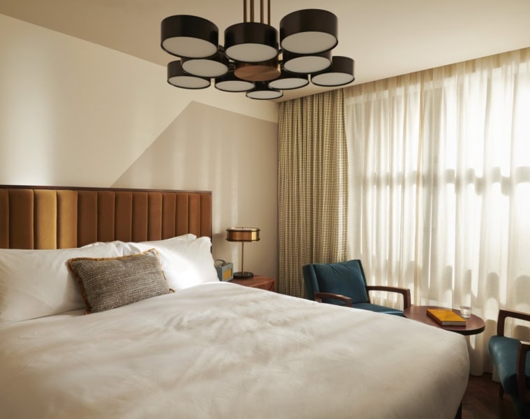 Ein Hotelzimmer im Mid-Century-Stil, mit breitem Bett, mit Samt gepolstertem Kopfteil und einer schwarzen Kunststoff-Designlampe an der Decke.