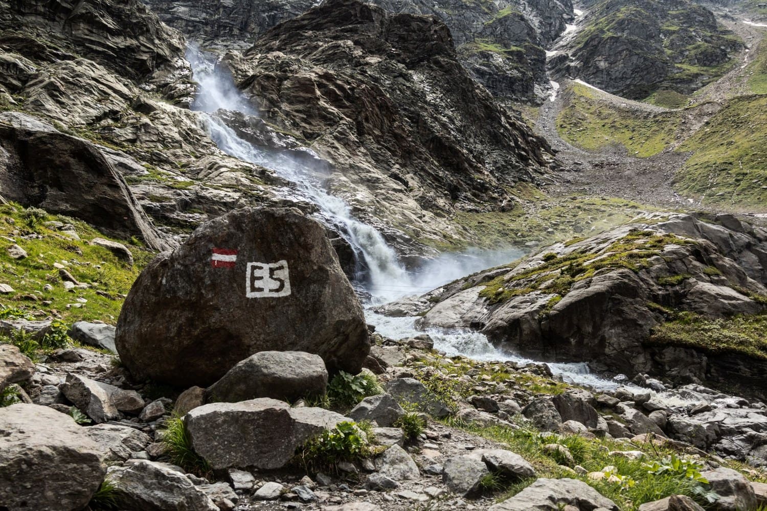 Felsblock mit rot-weiß-roter Wegmarkierung und E5-Schild vor einem Wasserfall in Tirol