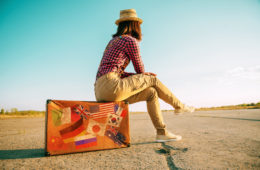 Weltreise: Frau mit langer Hose, Bluse und Hut sitzt auf einem Koffer an einer einsamen Landstraße