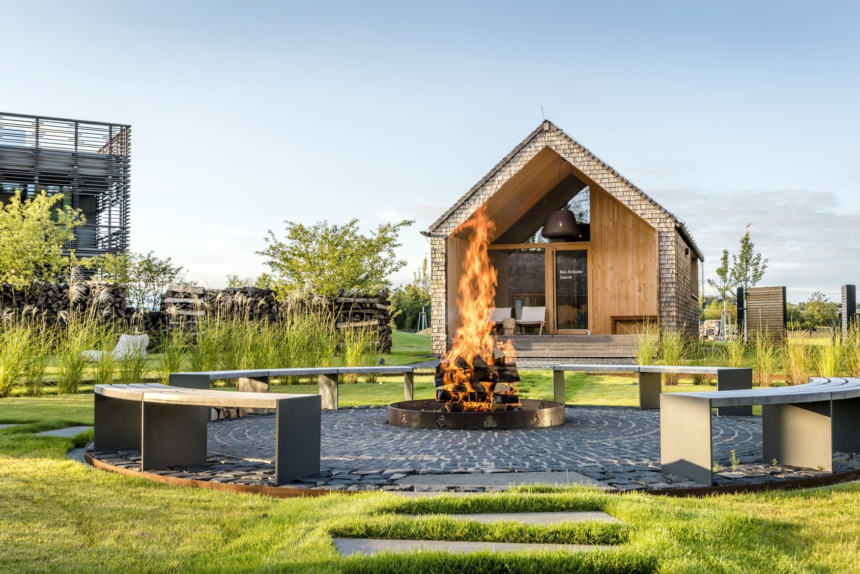Ein Feuer lodert in einer runden Feuerstelle auf gepflastertem Grund, ringsum geschwungene Bänke. Dahinter steht die Bio-Kräuter-Sauna mit Schindelfassade und offenem Giebel, links Holzstapel und Gräser.