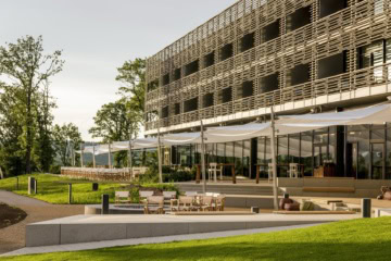 Hotelgebäude mit horizontaler Holzlamellenfassade im Abendlicht. Davor spannen sich weiße Sonnensegel über die Terrasse, auf den Stufen stehen Regiestühle um eine Feuerschale.