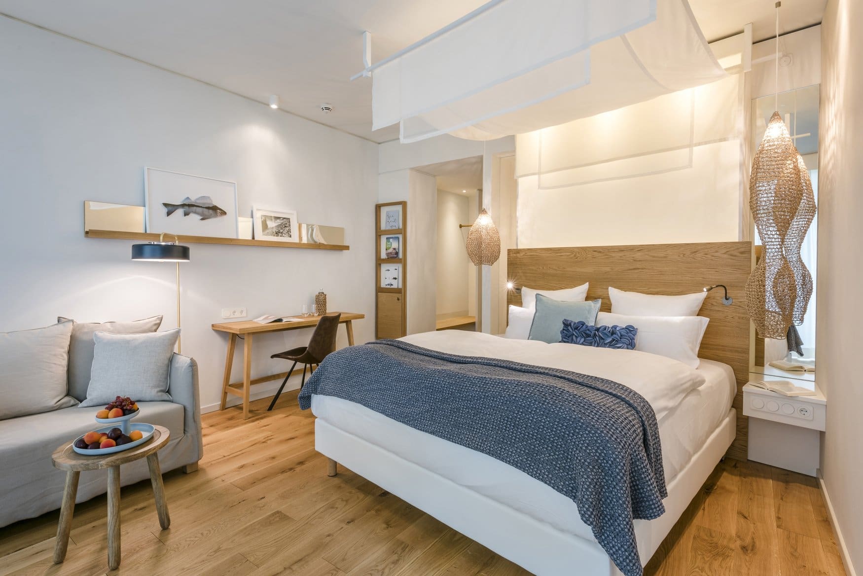 Hotelzimmer mit Doppelbett vor hölzernem Kopfteil, darüber ein Baldachin aus weißem Stoff. An der Wand hängt ein gerahmtes Fischbild über einem Wandbord, links steht ein Sofa, rechts eine geflochtene Hängeleuchte.