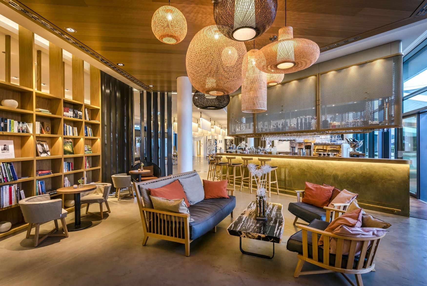 Lobbybar mit langem Tresen aus Messing und Barhockern, darüber hängen große geflochtene Leuchten. Links ein hohes Bücherregal, davor Sitzgruppen aus Holzsesseln mit blauen und orangefarbenen Kissen.