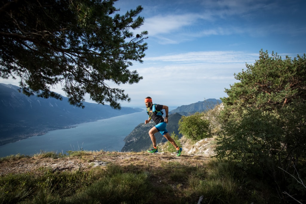 Sportlicher Mann joggt auf Bergpfad wischen Bäumen und Seeblick