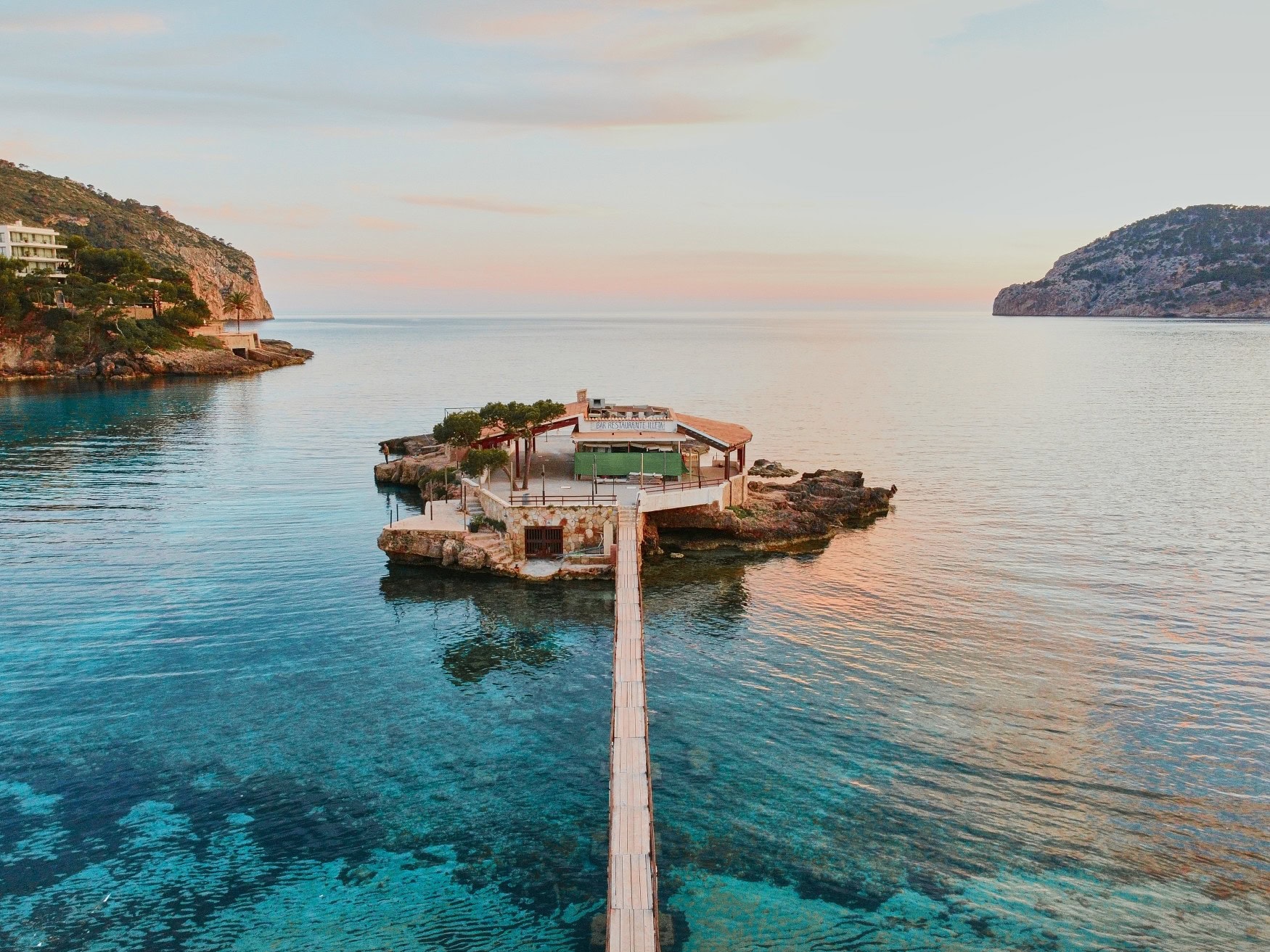 Ein hölzerner Steg führt durch das türkisblaue Wasser vor Mallorca über das Wasser bis zu einer kleinen Insel, darauf ein einzelnes Gebäude
