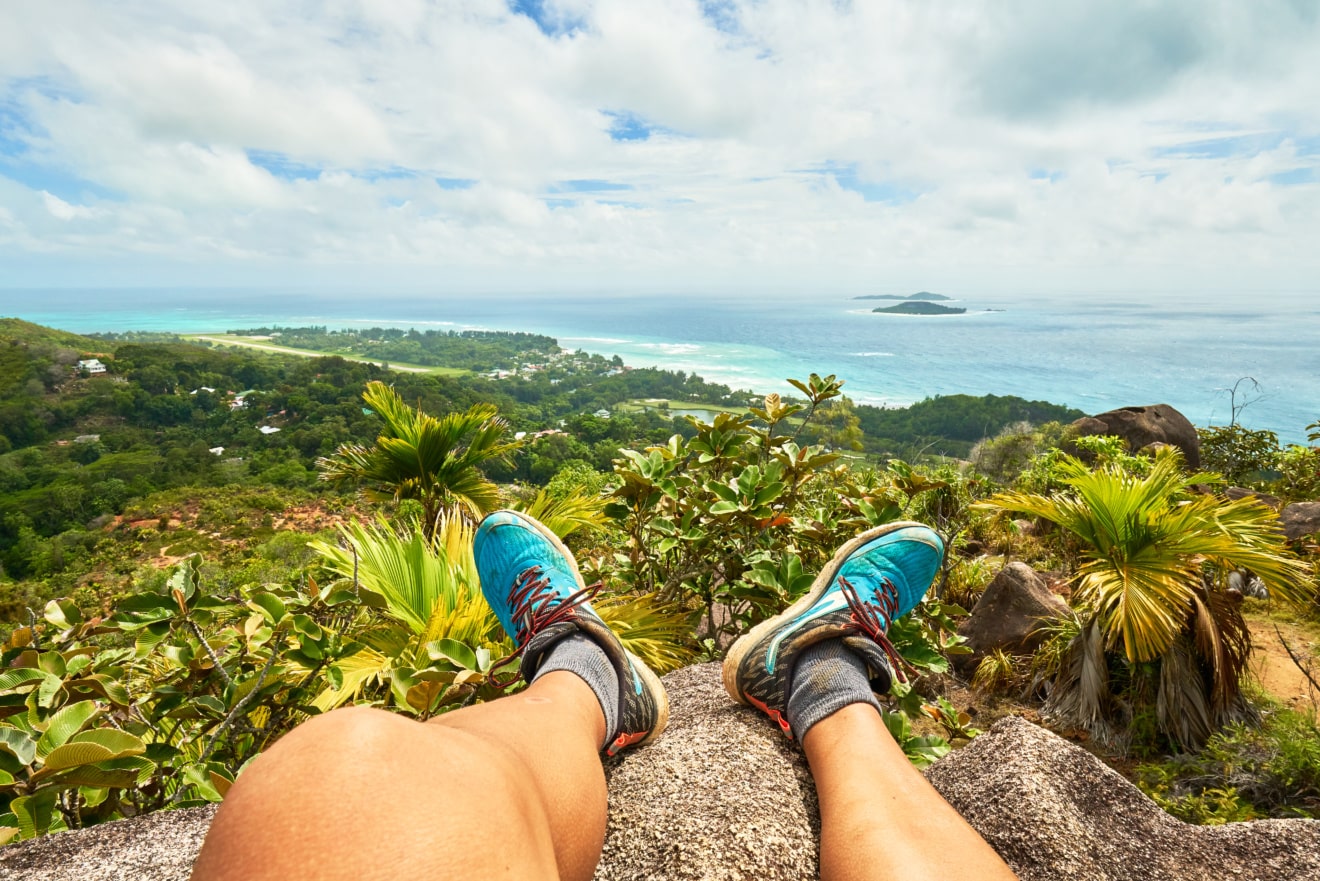 Wanderer mit ausgestreckten Beinen auf Plateau blickt auf die Küste der Seychellen