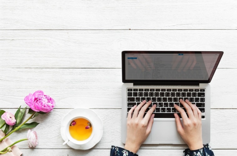 Person tippt auf Tastatur eines Notebooks, links davon eine Tasse Tee und eine rosafarbene Rose
