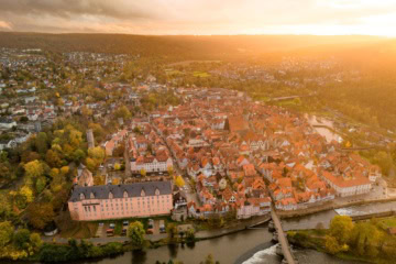 Drohnenansicht der Stadt Hann. Münden in Niedersachsen im Sonnenuntergang. Im Vordergrund ein Fluss mit einer Brücke darüber, mittig eine kleine Altstadt mit zahlreichen Fachwerkhäusern, deren rot Dächer im Licht der Sonne strahlen.