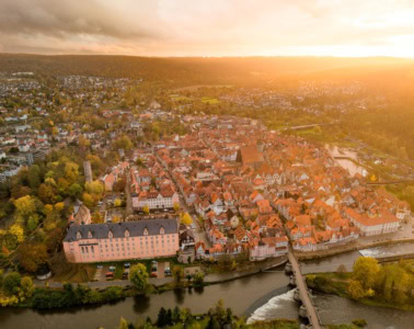 Drohnenansicht der Stadt Hann. Münden in Niedersachsen im Sonnenuntergang. Im Vordergrund ein Fluss mit einer Brücke darüber, mittig eine kleine Altstadt mit zahlreichen Fachwerkhäusern, deren rot Dächer im Licht der Sonne strahlen.