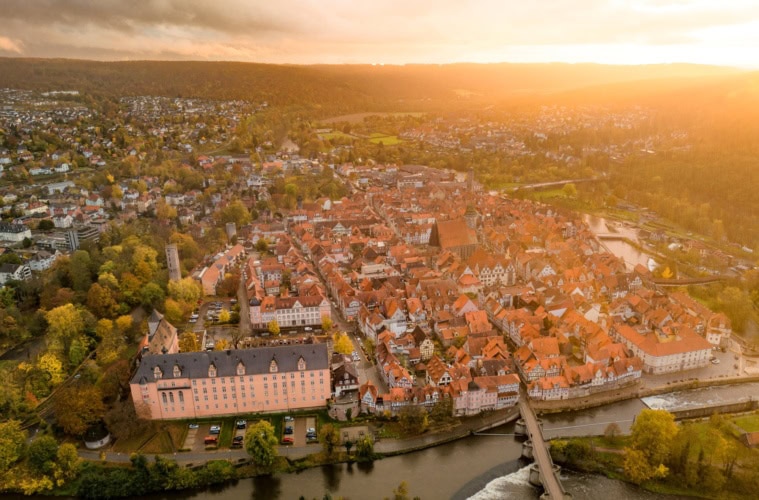 Drohnenansicht der Stadt Hann. Münden in Niedersachsen im Sonnenuntergang. Im Vordergrund ein Fluss mit einer Brücke darüber, mittig eine kleine Altstadt mit zahlreichen Fachwerkhäusern, deren rot Dächer im Licht der Sonne strahlen.