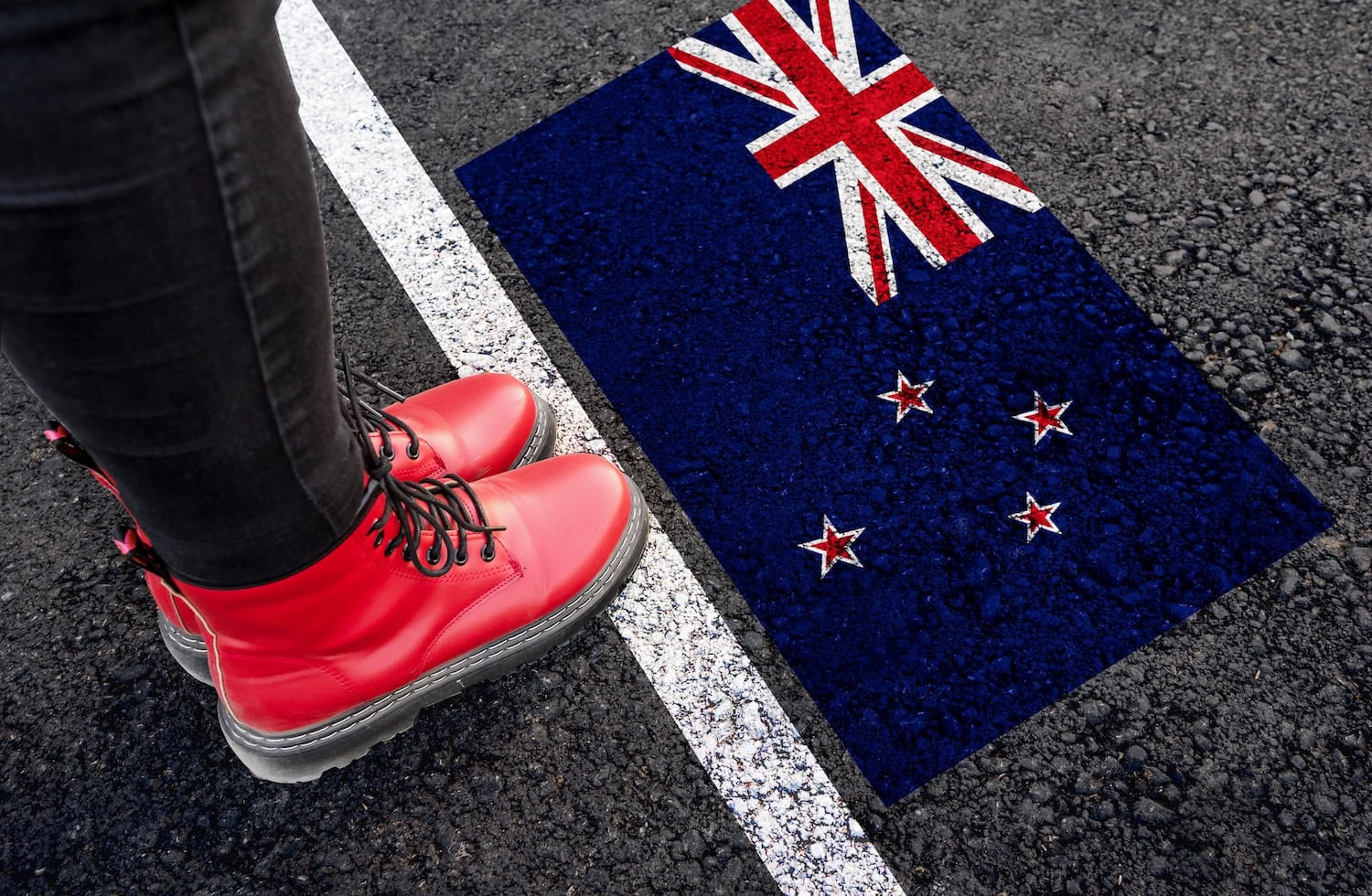 Eine Frau in Stiefeln steht auf Asphalt neben der Flagge Neuseelands und einer Grenzmarkierung.