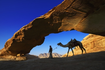 Ein Beduin reitet sein Kamel durch den Steinbogen "Al Kharza" im Wadi Rum, Jordanien.