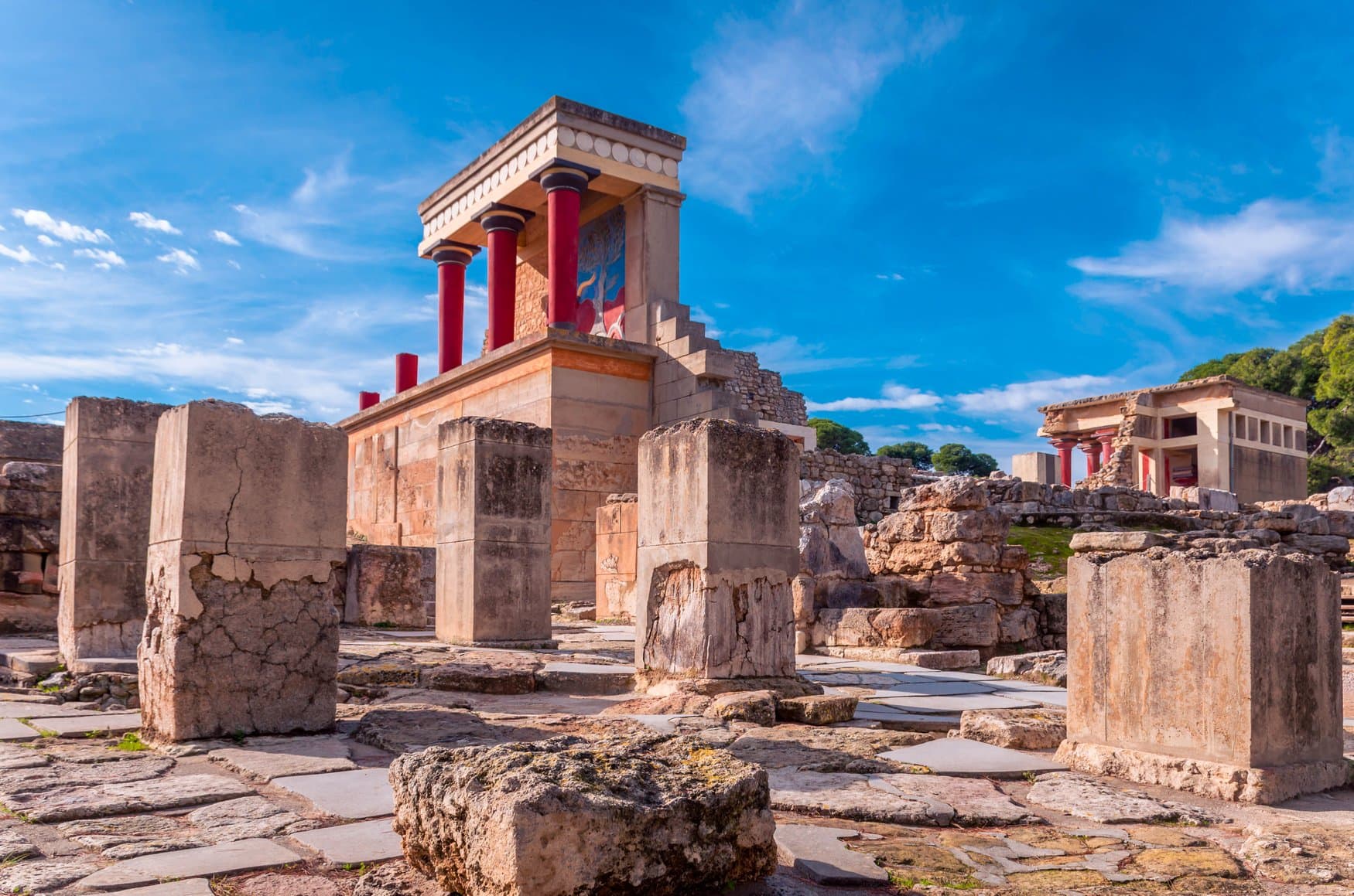 Rekonstruierter Teil des minoischen Palasts von Knossos auf Kreta mit den markanten rot gestrichenen Säulen und einem Stierfresko, umgeben von Mauerresten unter blauem Himmel.