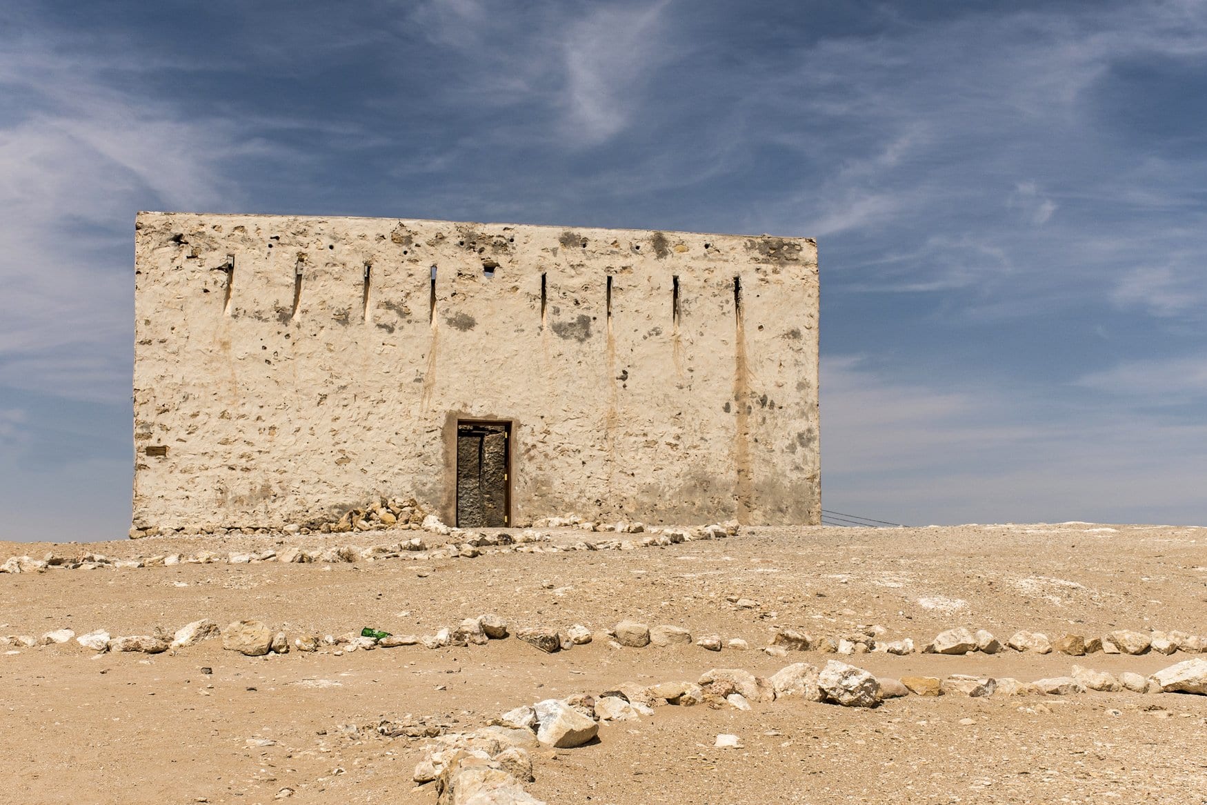 Freistehendes verwittertes Steingebäude mit heller Verputzung und schmaler Tür in der Wüste des Oman, davor lose Steine unter blauem Himmel.