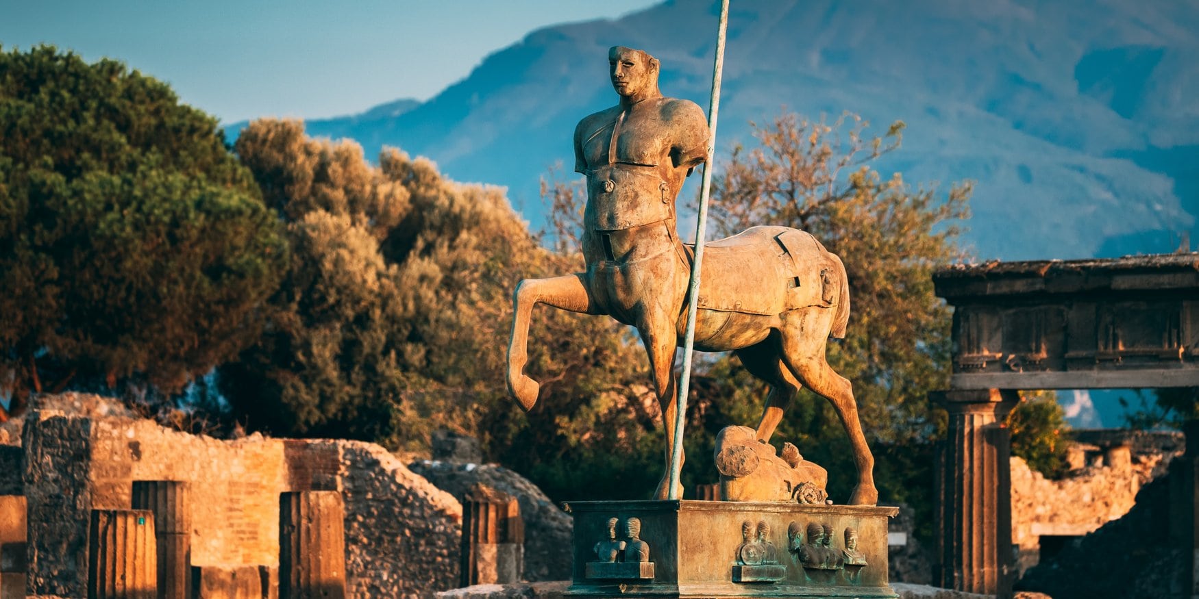 Bronzene Zentaurenstatue im antiken Pompeji, ein Mischwesen aus Mann und Pferd auf einem Sockel, dahinter Ruinen und der Vesuv im Abendlicht.