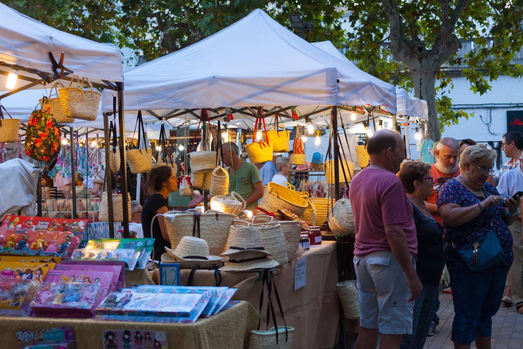 Marktstände mit Körben, Taschen und Strohhüten unter weißen Pavillons beim abendlichen Markt in Maria de la Salut, davor Besucher am Verkaufstisch.