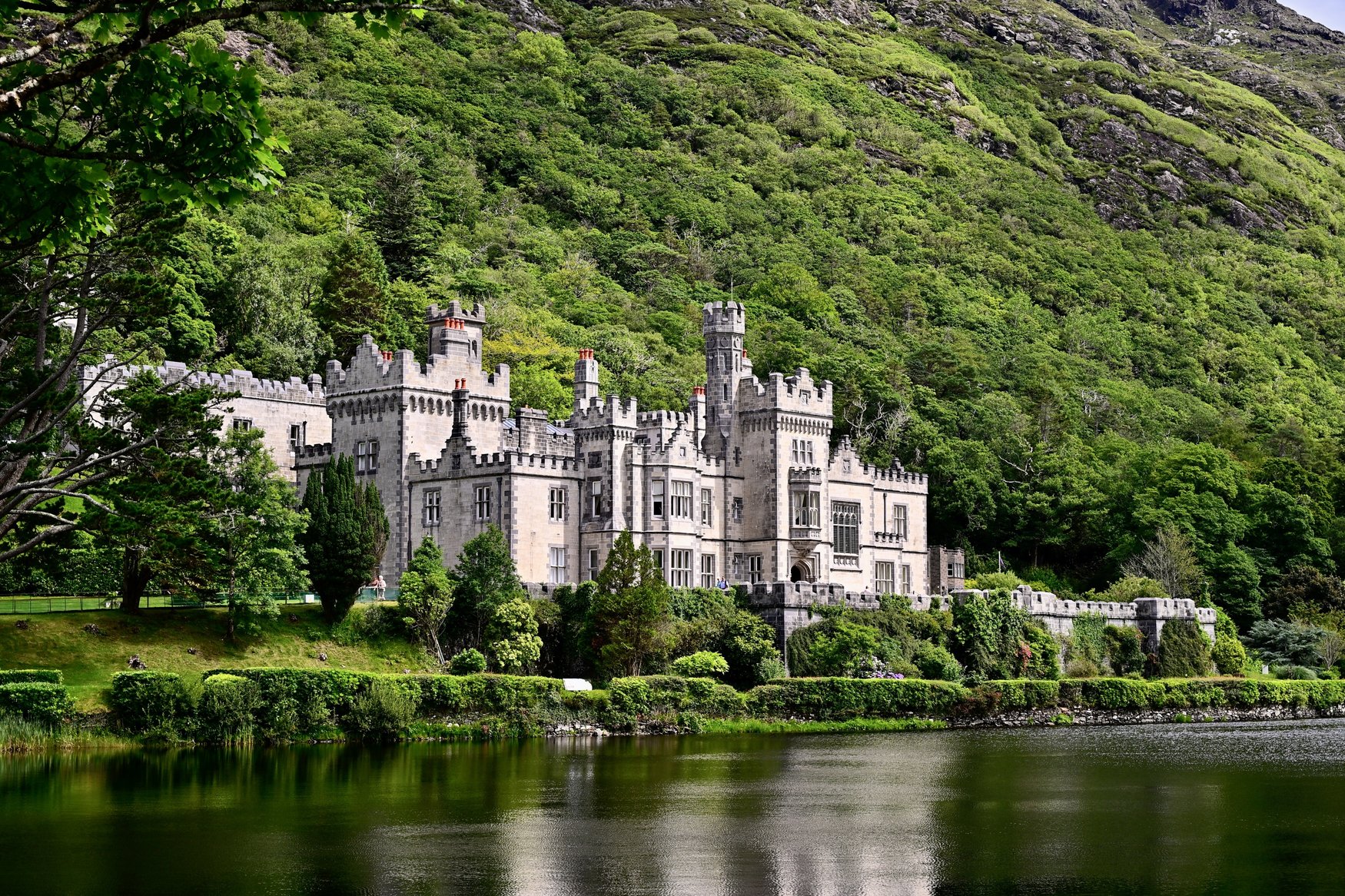 Kylemore Abbey mit Zinnen und Türmen am Seeufer, dahinter steigt ein dicht bewachsener Hang an.