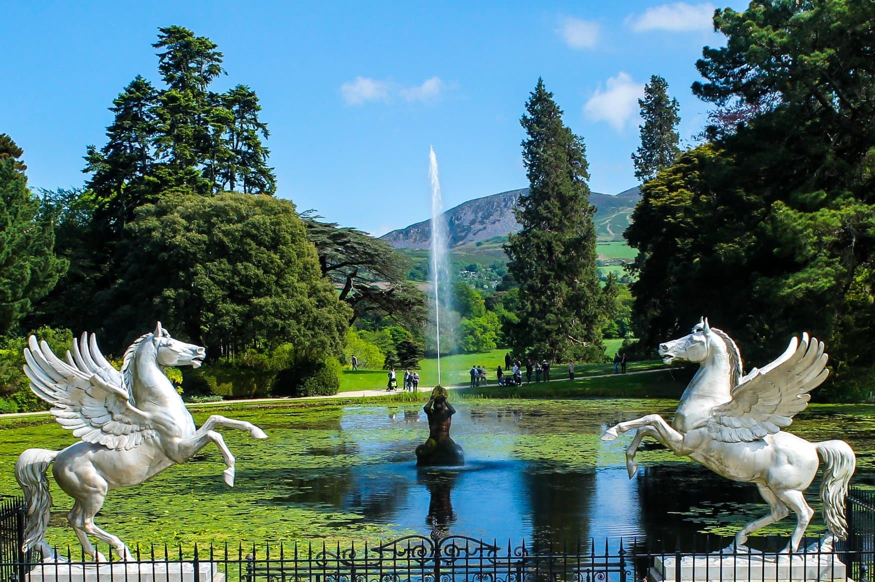 Zwei weiße Pegasusfiguren flankieren das Wasserbecken mit hoher Fontäne in den Powerscourt Gardens, dahinter Bäume und Hügel.