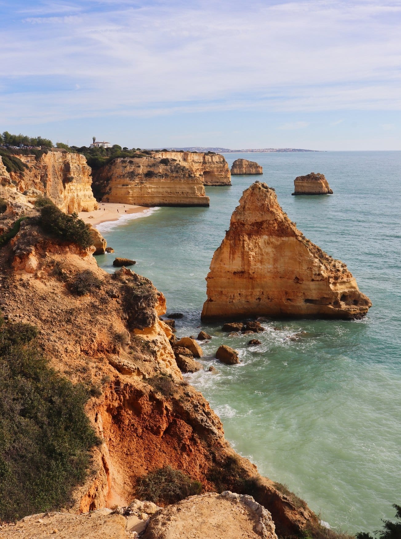 Blick entlang der Algarveküste auf eine Bucht mit hellem Sandstrand. Im Wasser stehen mehrere freistehende Felstürme, auf der Klippe links liegt ein weißes Gebäude zwischen Bäumen.