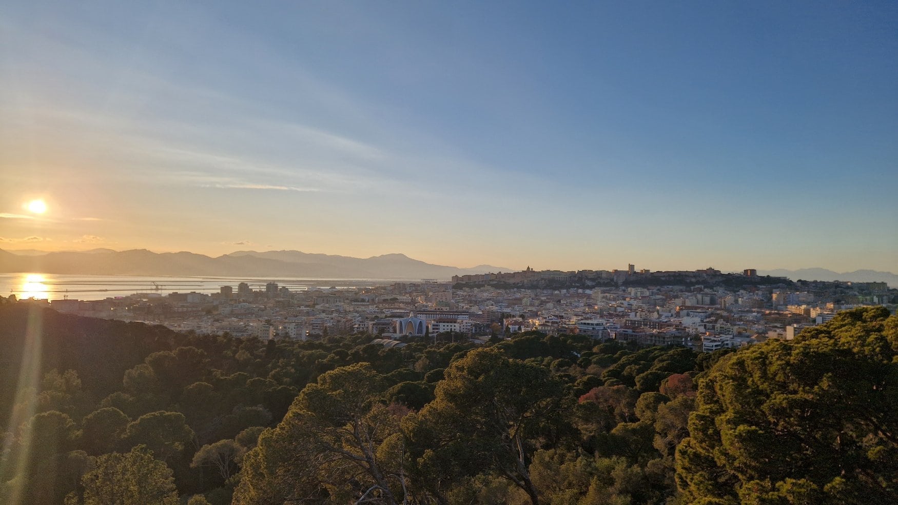 Blick von einer bewaldeten Anhöhe über Cagliari auf Sardinien bei Sonnenuntergang. Hinter den Dächern der Stadt liegen der Hafen und die Lagune, am Horizont stehen die Berge im Dunst.