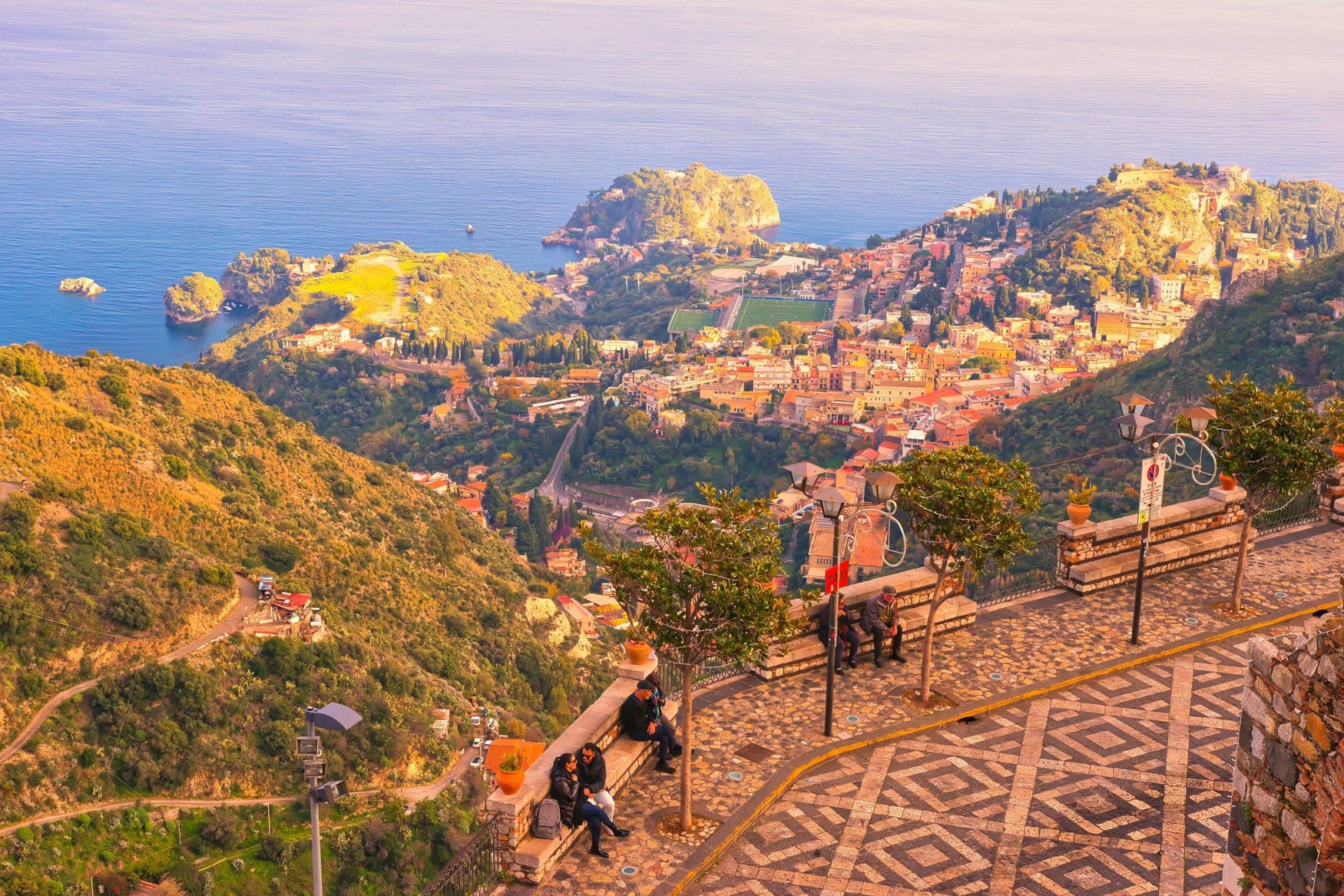 Blick von der Aussichtsterrasse in Castelmola auf Taormina und die Küste Siziliens. Im Vordergrund sitzen Besucher auf einer Mauer zwischen Bäumen und Laternen, der Platz ist mit einem Rautenmuster gepflastert.