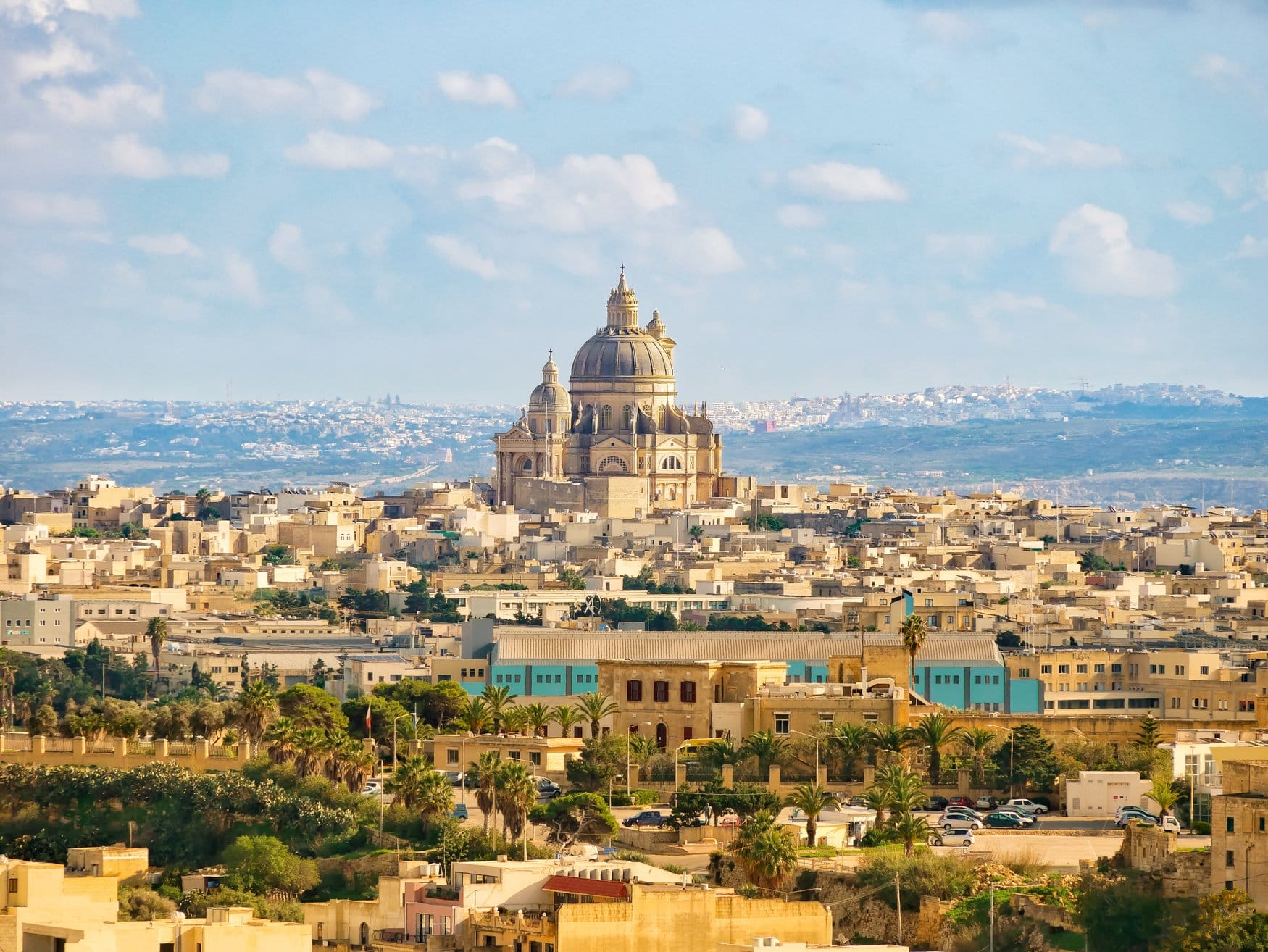 Blick über eine Ortschaft aus hellem Kalkstein auf Malta. Über den Flachdächern erhebt sich eine Kirche mit mächtiger Kuppel und Laterne, im Vordergrund stehen Palmen und Zypressen.