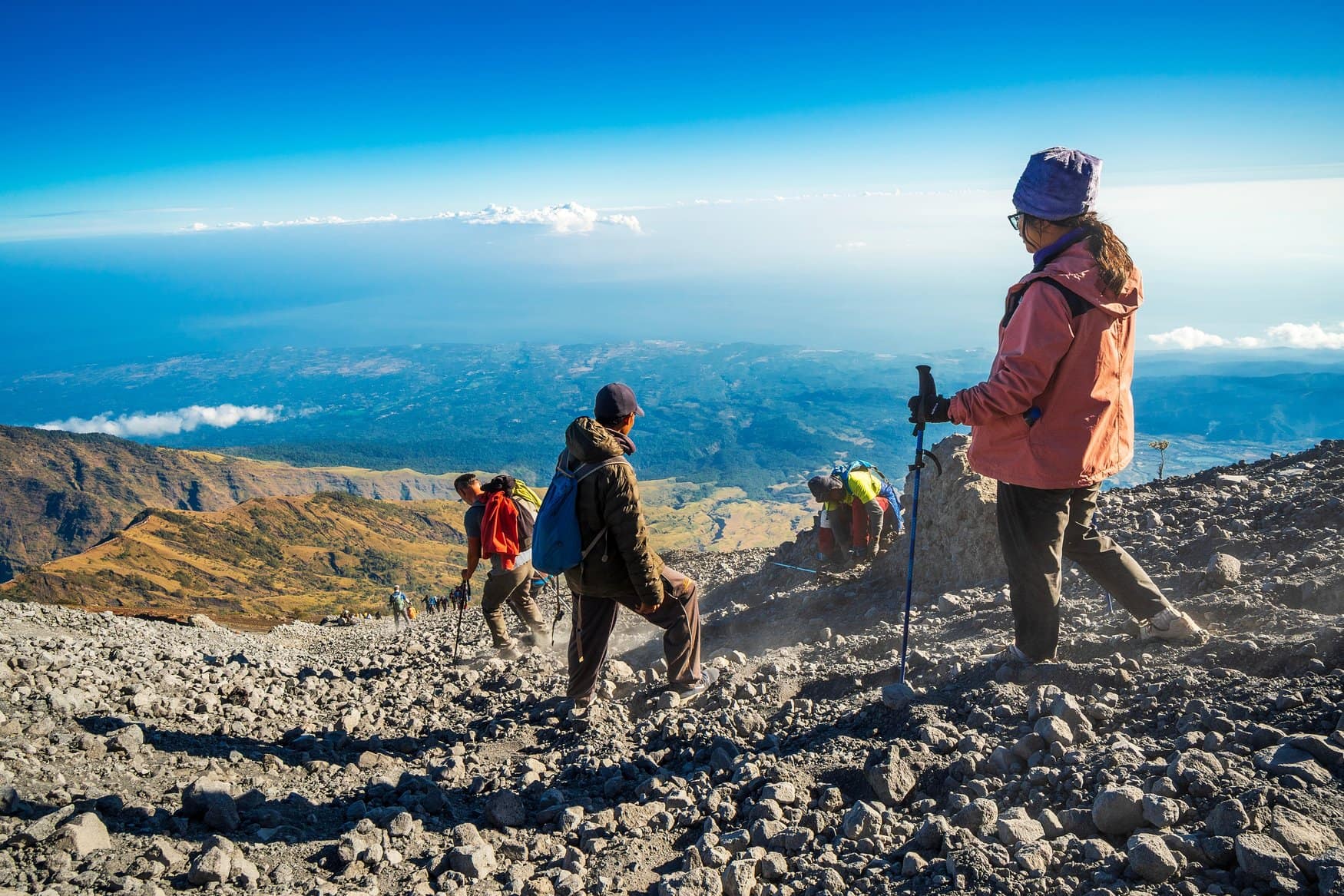 Wanderer und Träger steigen über ein Geröllfeld am Mount Rinjani auf Lombok ab, im Hintergrund öffnet sich der Blick über die Kraterwand auf das Tiefland und das Meer.