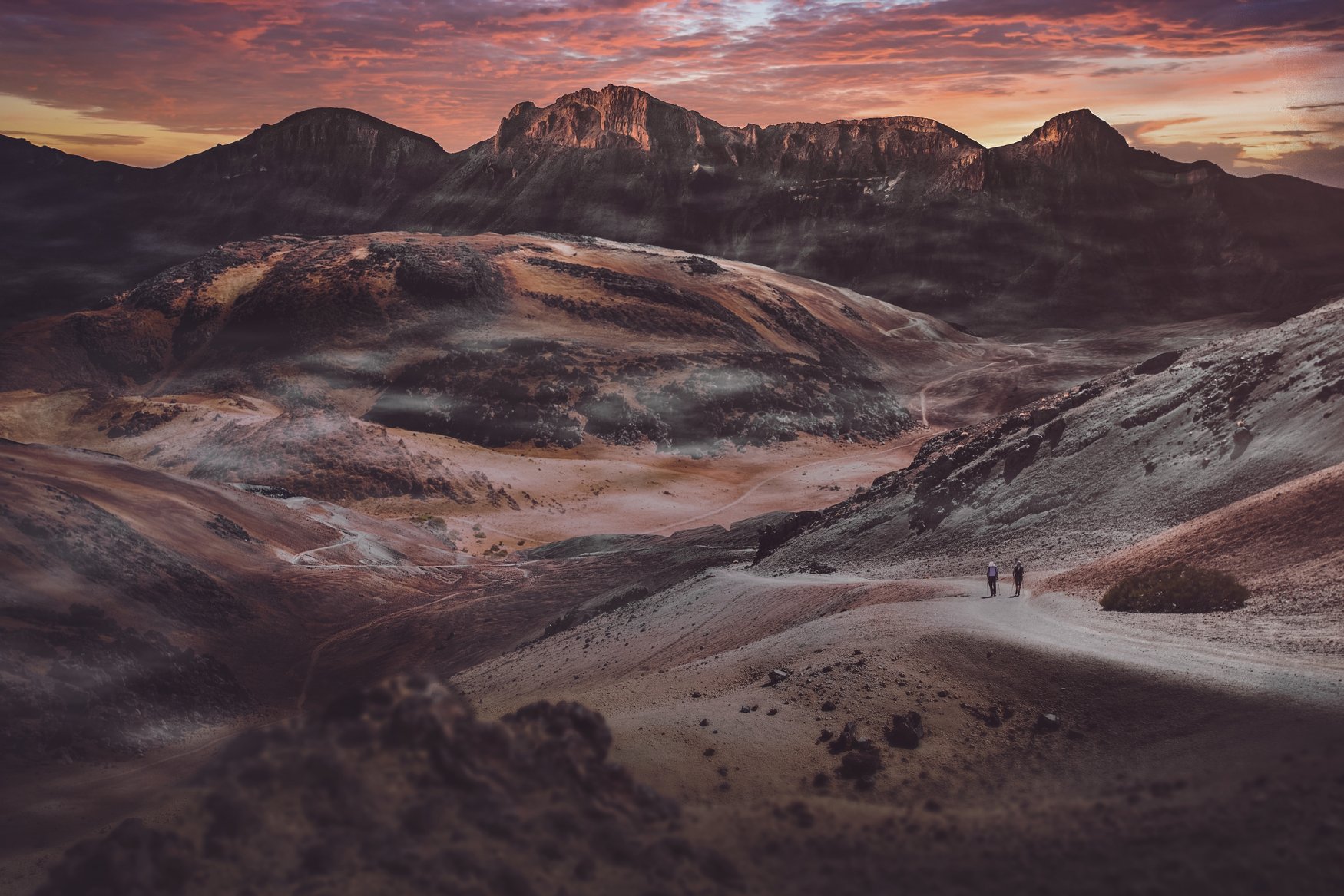 Zwei Wanderer gehen auf einem Schotterweg durch die helle Vulkanlandschaft des Teide-Nationalparks. Dahinter steigt ein zerklüfteter Bergkamm auf, der Himmel ist rot gefärbt.