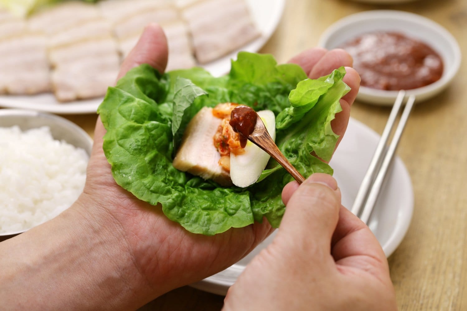 Eine Hand hält ein Salatblatt mit einer Scheibe gekochtem Schweinebauch und Kimchi, die andere gibt mit einem Holzlöffel würzige Ssamjang-Paste darauf; im Hintergrund stehen eine Platte mit aufgeschnittenem Bossam, eine Schale Reis und ein Schälchen mit Sauce.