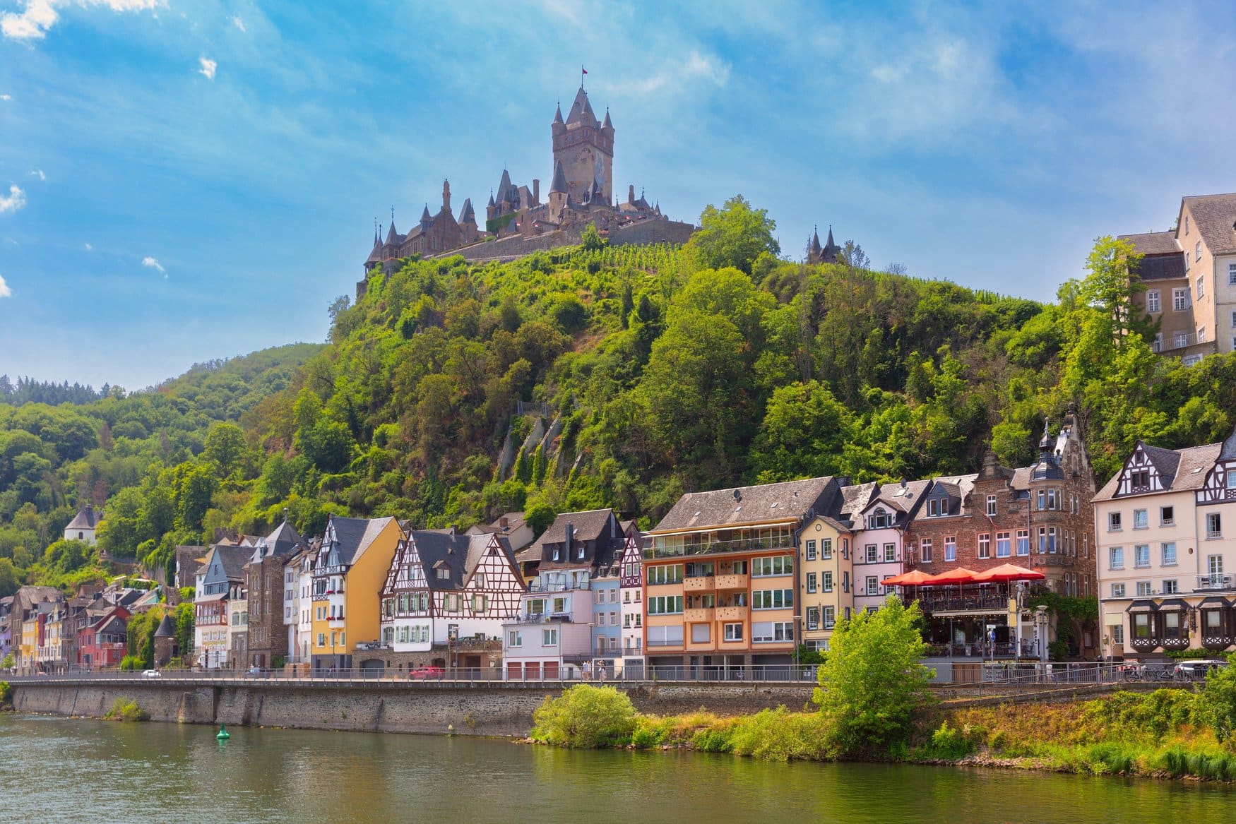 Die Reichsburg Cochem auf ihrem bewaldeten Bergkegel über der Mosel. Am Ufer stehen bunte Fachwerkhäuser hinter der Uferpromenade.