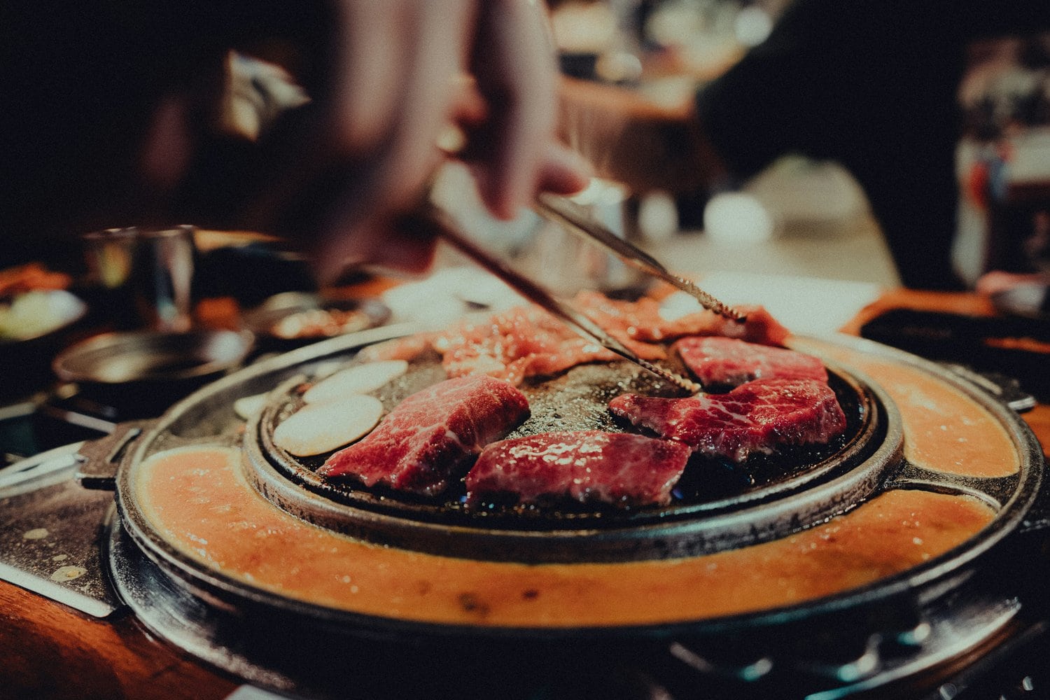 Stark marmorierte Rindfleischstücke und Knoblauchscheiben garen auf einem runden Tischgrill in einem koreanischen BBQ-Restaurant, eine Hand wendet das Fleisch mit einer Grillzange, im Ring um die Grillplatte stockt eine Ei-Masse.