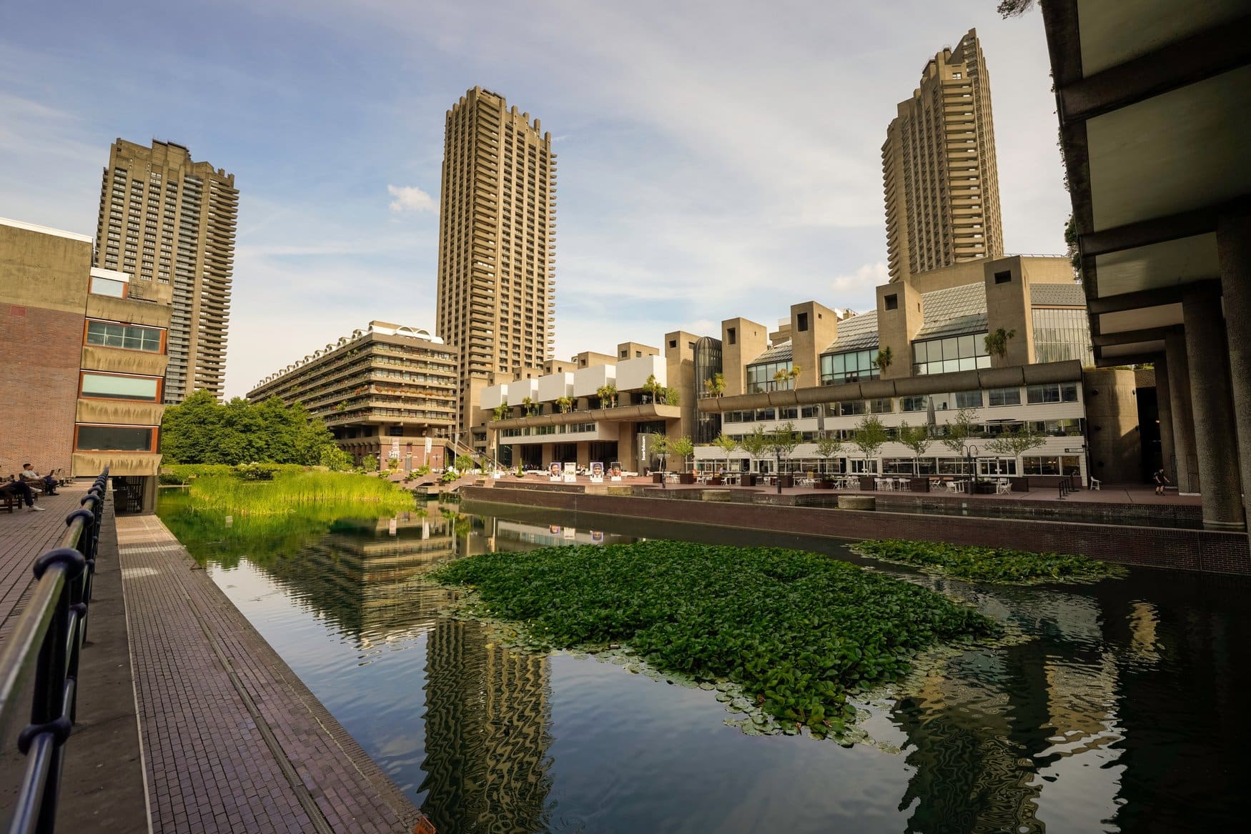 Das Barbican Estate in London mit den Betontürmen der Wohnanlage und dem Barbican Centre. Im Vordergrund liegt ein Wasserbecken mit Seerosen, das die Gebäude spiegelt.