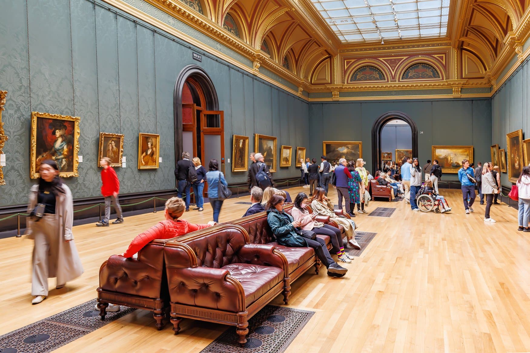 Ausstellungssaal der National Gallery in London mit grün tapezierten Wänden und vergoldeter Deckenkassettierung. Besucher betrachten die Gemälde, in der Mitte steht eine lederne Sitzbank.