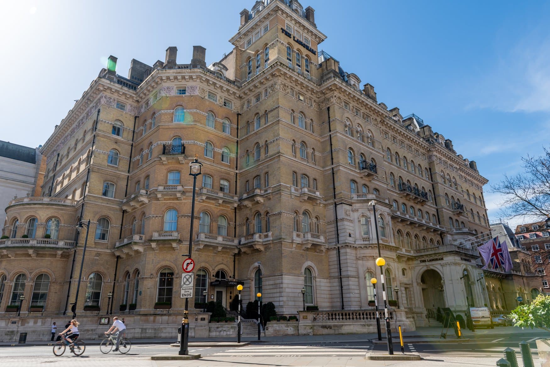 Die Fassade des Hotels The Langham an der Regent Street in London. Vor dem Sandsteinbau fahren zwei Radfahrer über die Straße, am Eingang hängt eine Union Jack.