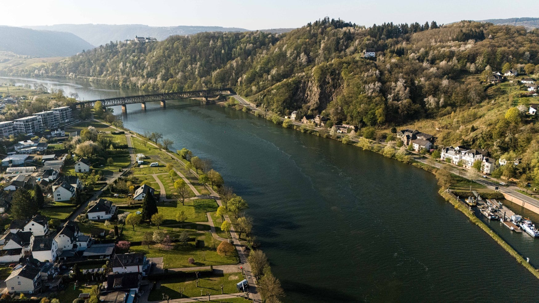 Luftaufnahme einer Moselschleife mit einer stählernen Eisenbahnbrücke. Am linken Ufer liegt ein Ort mit Uferpark, rechts steigt ein bewaldeter Hang mit blühenden Bäumen an.