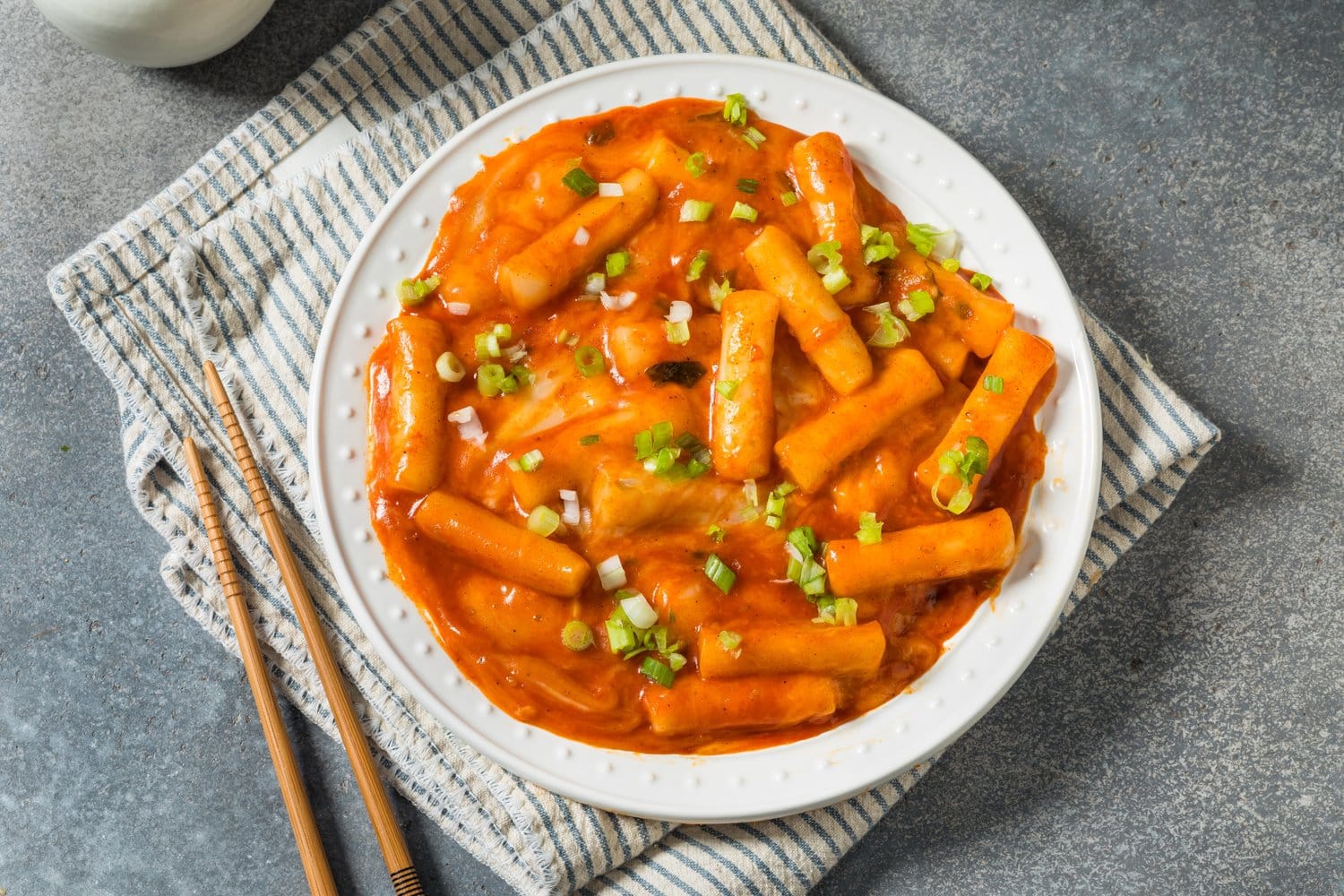 Tteokbokki, koreanische Reiskuchen-Stangen in cremiger, orangeroter Chilisauce mit geschmolzenem Käse, bestreut mit Frühlingszwiebeln, auf einem weißen Teller auf einem gestreiften Leinentuch, daneben Essstäbchen aus Holz.