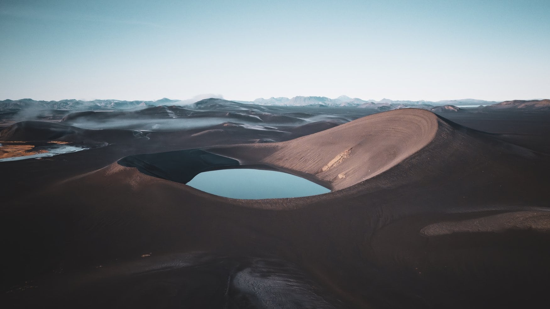 Luftaufnahme einer schwarzen Vulkanlandschaft auf Island mit einem türkisfarbenen Kratersee, der in einer Senke aus dunkler Vulkanasche liegt, im Hintergrund flache Berge am Horizont.