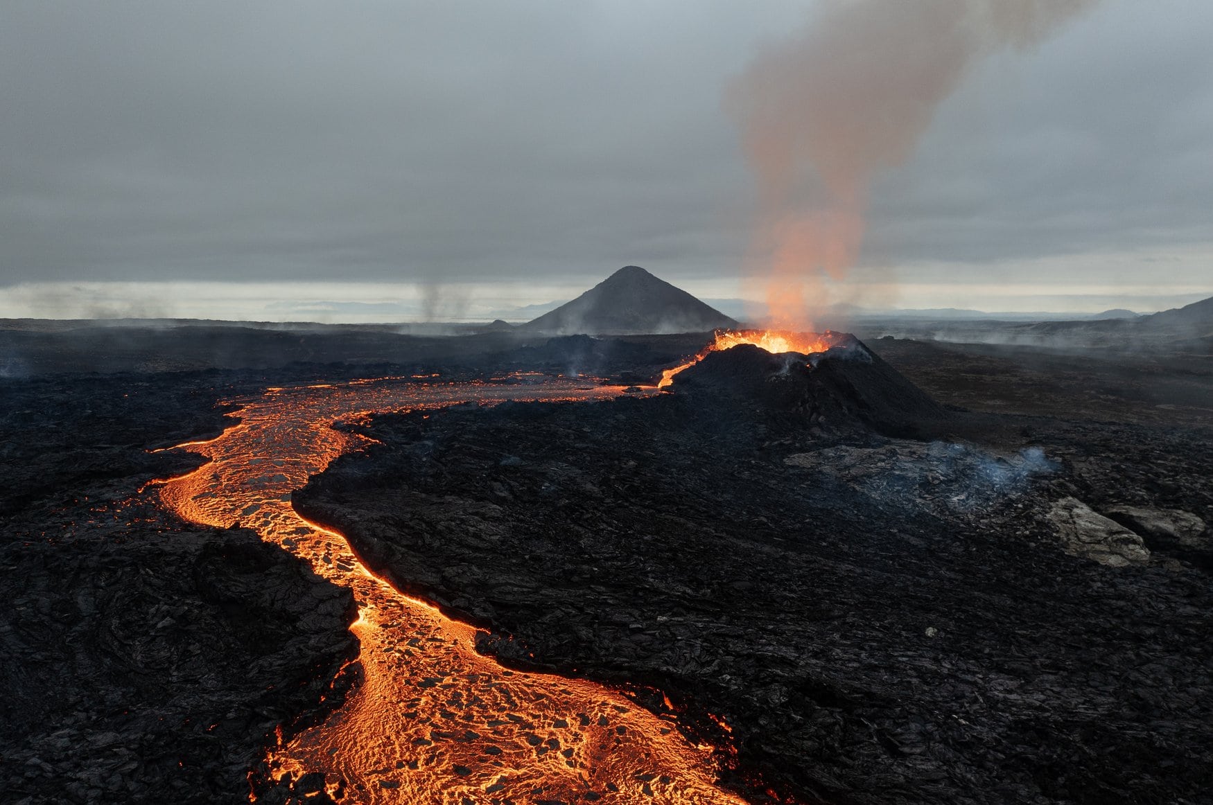 Vulkanausbruch auf Island mit einem aktiven Kraterkegel, aus dem Lava spritzt und eine Rauchsäule aufsteigt, davor ein glühender Lavastrom durch ein schwarzes Lavafeld unter grauem Himmel.