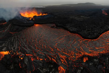 Vulkanausbruch auf Island mit einem aktiven Kraterkegel, aus dem Lava spritzt und eine Rauchsäule aufsteigt, davor ein glühender Lavastrom durch ein schwarzes Lavafeld unter grauem Himmel.