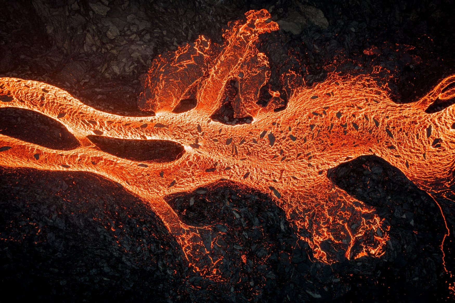 Nahaufnahme aus der Luft von glühender, orange leuchtender Lava, die sich in verzweigten Strömen über bereits erkaltetes schwarzes Gestein bewegt.