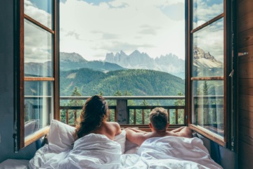 Pärchen liegt im Bett und blickt aus offenem Fenster auf die umliegende Bergkulisse