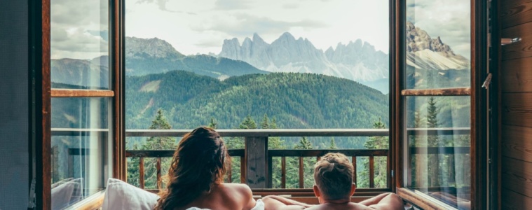 Pärchen liegt im Bett und blickt aus offenem Fenster auf die umliegende Bergkulisse