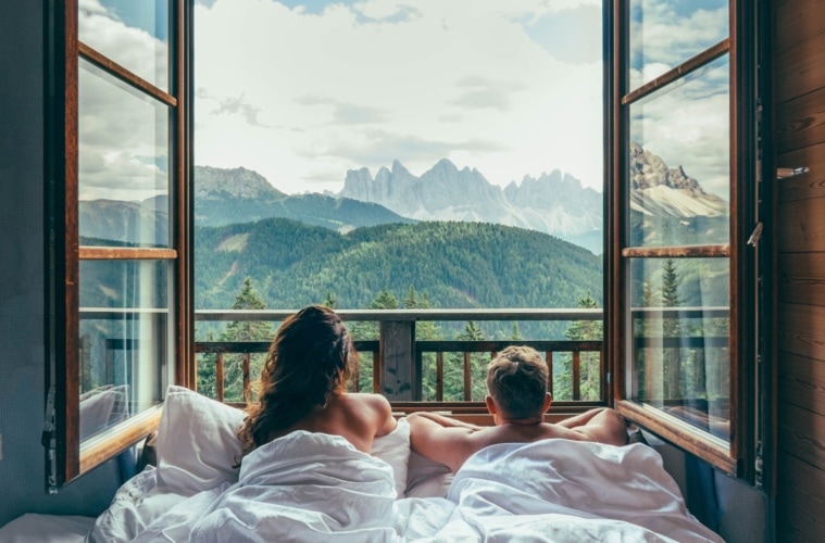 Pärchen liegt im Bett und blickt aus offenem Fenster auf die umliegende Bergkulisse